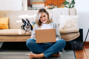 A blond, middle-aged woman sitting cross legged on the floor of her living room researching on her laptop. Her dog, a Boston Terrier, sits on the sofa behind her.