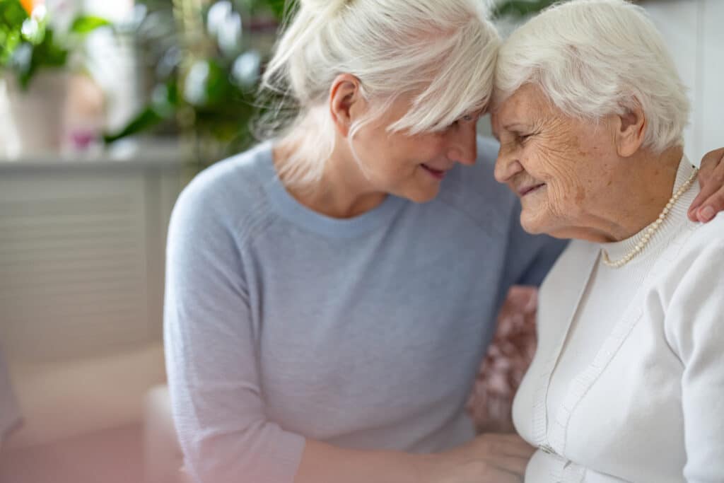 An adult daughter with white hair pulled up, wearing a blue shirt leaning lovingly in to put her forehead against her elderly mother's. Her mother has short white hair and is wearing a white shirt. They have an obvious familial resemblance.